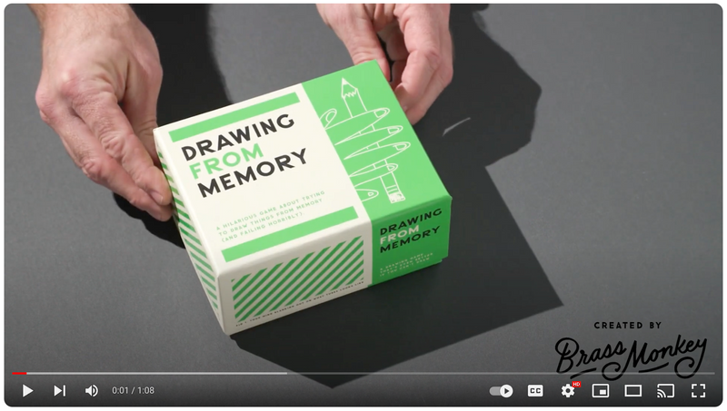 A person’s hands hold a green and white box labeled “Drawing From Memory.” The box features simple pencil illustrations and is set against a dark surface. The bottom right shows the text “Created by Brass Monkey.”.