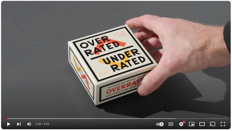 A hand is reaching for a square game box labeled OVERRATED UNDERRATED on a dark surface. The box features bold, colorful text and geometric design elements.