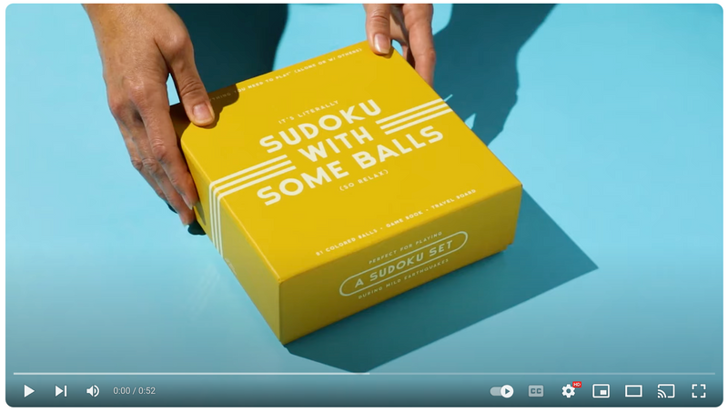 A person’s hands holding a bright yellow box on a blue background. The box reads Its literally Sudoku with some balls and A Sudoku Set in bold white text.