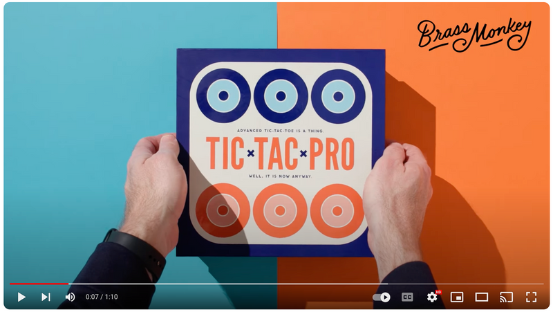 A person holds a square game box labeled Tic Tac Pro with circular designs on a split orange and blue background. The Brass Monkey logo is in the top right corner.