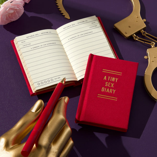 A red Brass Monkey A Tiny Sex Diary rests beside an open log page, a matching red pen, gold handcuffs, a gold mannequin hand, and a pink rose on a purple surface.