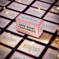 A pink Brass Monkey Date Night Challenges box sits among unique challenge cards, with warm fairy lights adding a cozy touch—creating the perfect, inviting atmosphere for memorable date nights.