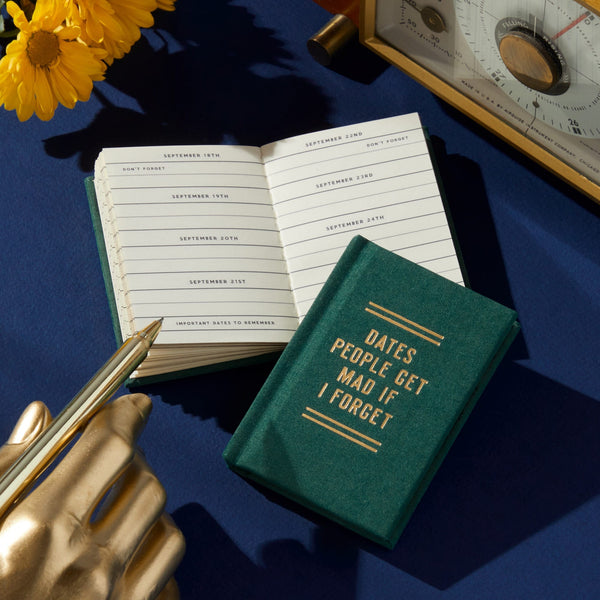 A green, hard-bound Dates People Get Mad If I Forget Tiny Diary by Brass Monkey lies open on a blue surface beside a gold hand-shaped pen holder, pen, yellow flowers, and a vintage clock.