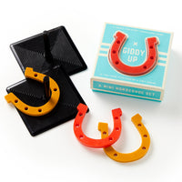 The Brass Monkey Giddy Up Tabletop Horseshoes Game includes a blue box, two black bases with pegs, and four small rubber horseshoes—two red and two yellow—displayed on a white background.