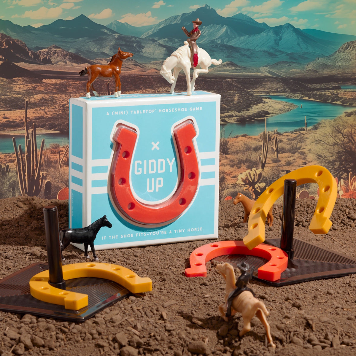 The Brass Monkey Giddy Up Tabletop Horseshoes Game is shown in a desert scene with toy horses, cowboys, rubber horseshoes, and game pieces arranged on brown soil before mountains and a river.
