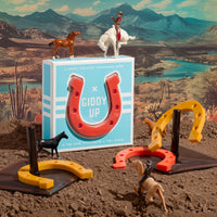 The Brass Monkey Giddy Up Tabletop Horseshoes Game is shown in a desert scene with toy horses, cowboys, rubber horseshoes, and game pieces arranged on brown soil before mountains and a river.