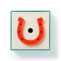 A bright red rubber horseshoe from the Brass Monkey Giddy Up Tabletop Horseshoes Game sits atop its box, which promises all game pieces and instructions are tucked inside—perfect for an indoor party match.