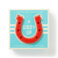 Brass Monkey’s Giddy Up Tabletop Horseshoes Game comes in a box with a red horseshoe on a blue and white background. Text reads: “A (mini) tabletop indoor horseshoes party game. If the shoe fits—you’re a tiny horse.”.