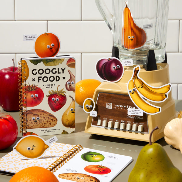 A playful scene features fruits and kitchen items with googly eyes, including a smiling blender with an onion, Brass Monkey stickers, the Googly Food Sticker Book by Brass Monkey, and notebooks—all with cute googly-eyed faces.