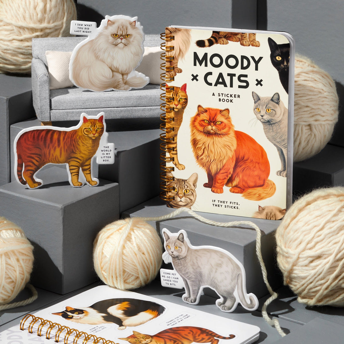 The Brass Monkey Moody Cats Sticker Book, featuring illustrated cats, is displayed with vintage-style puffy stickers, tiny couches, and large cream yarn balls on gray platforms.