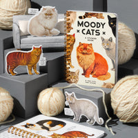 The Brass Monkey Moody Cats Sticker Book, featuring illustrated cats, is displayed with vintage-style puffy stickers, tiny couches, and large cream yarn balls on gray platforms.