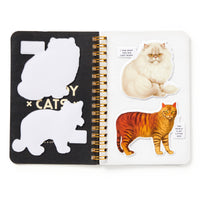 An open spiral notebook shows two Moody Cats stickers from Brass Monkey’s Moody Cats Sticker Book—a fluffy white cat and a striped brown cat—on the right page, with their empty outlines and playful speech bubbles on the left.