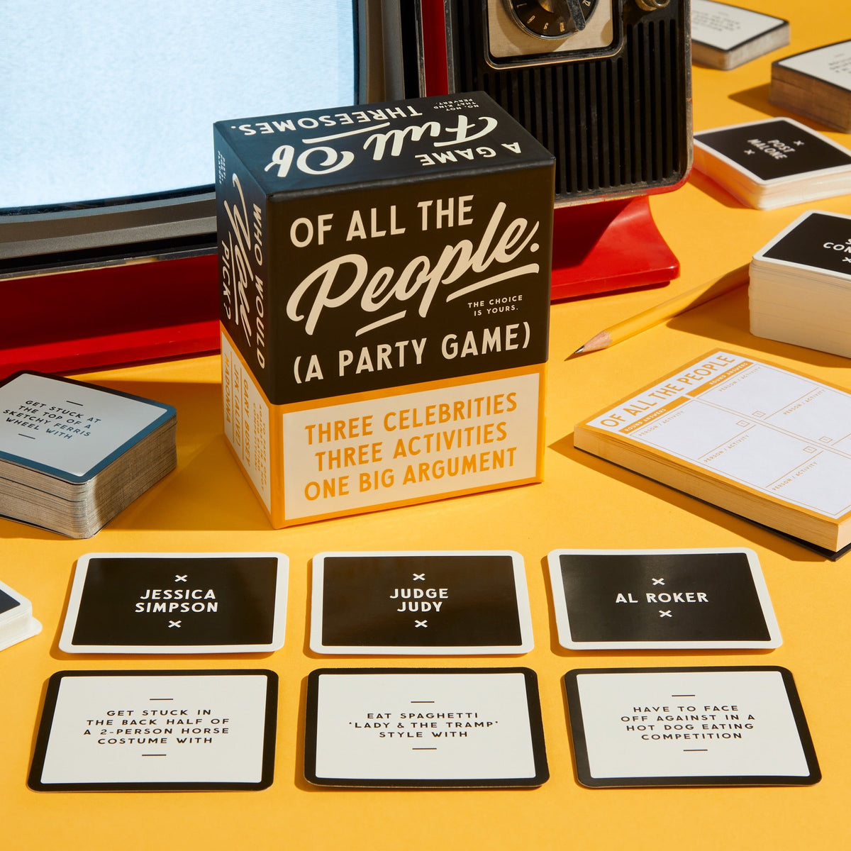 A Brass Monkey Of All The People Social Game box rests on a yellow surface with cards featuring celebrity names and activities spread around, while a vintage TV and notebook appear in the background.