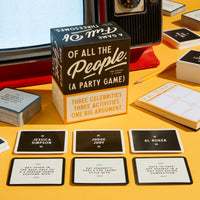 A Brass Monkey Of All The People Social Game box rests on a yellow surface with cards featuring celebrity names and activities spread around, while a vintage TV and notebook appear in the background.