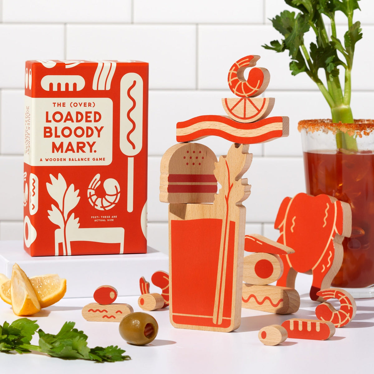 The (Over) Loaded Bloody Mary Balance Game by Brass Monkey sits next to wooden celery, lemon, and olive pieces stacked in a glass shape on a white counter with a tiled wall background.