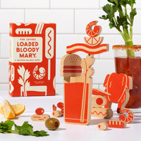 The (Over) Loaded Bloody Mary Balance Game by Brass Monkey sits next to wooden celery, lemon, and olive pieces stacked in a glass shape on a white counter with a tiled wall background.