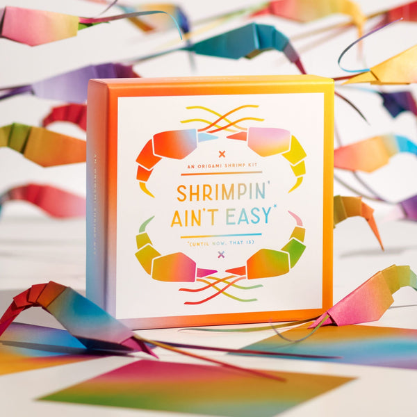 The Brass Monkey Shrimpin Aint Easy Origami Kit comes in a colorful box with rainbow shrimp artwork. Matching paper shrimp decorations surround the kit, emphasizing its playful origami theme on a white background.