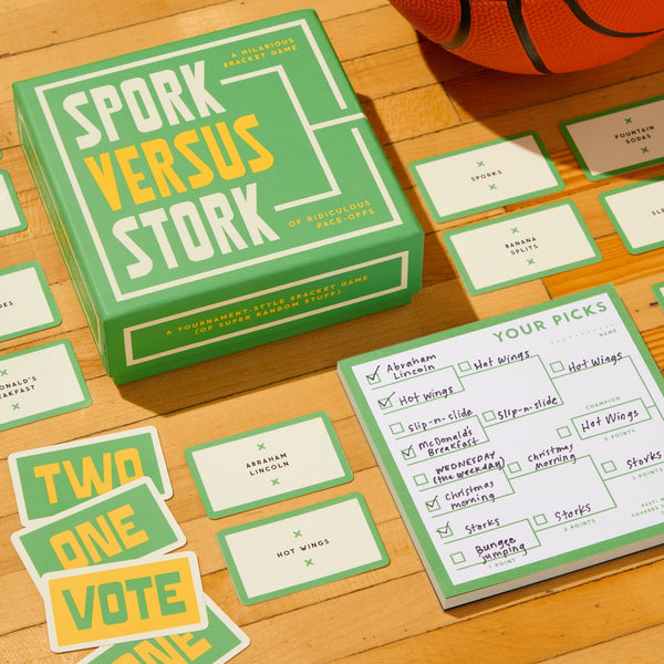 The Brass Monkey Spork Versus Stork Social Game is shown on a wooden surface with player cards, a scoring sheet, and part of a basketball. The cards display categories, numbers, and choices like Abraham Lincoln or Hot wings.