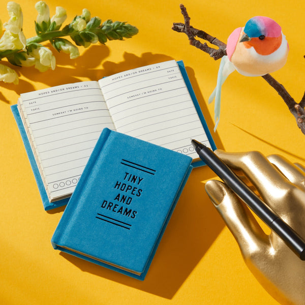 Brass Monkey’s Tiny Hopes and Dreams Tiny Diary, a small blue pocket journal, rests on a yellow surface beside an open page, a black pen in a gold hand holder, a flower, and a colorful felt bird on a branch.