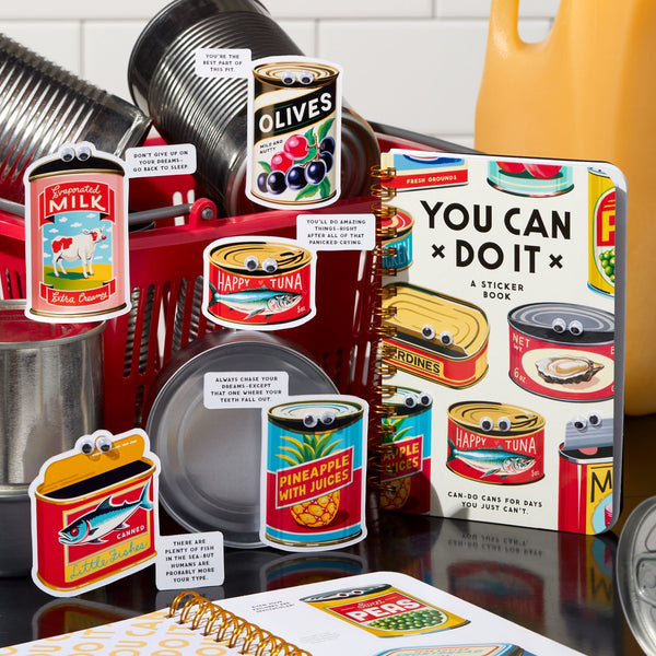 A collection of Brass Monkeys googly-eyed cans with colorful labels and motivational signs is displayed in a kitchen with dishes and utensils. In the center, the You Can Do It Googly Sticker Book stands out.