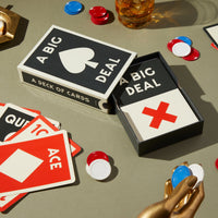 A Big Deal Giant Playing Cards by Brass Monkey are displayed on a table with some cards spread out, poker chips, and a drink. The oversized cards feature bold red and black geometric shapes and vintage-inspired designs.