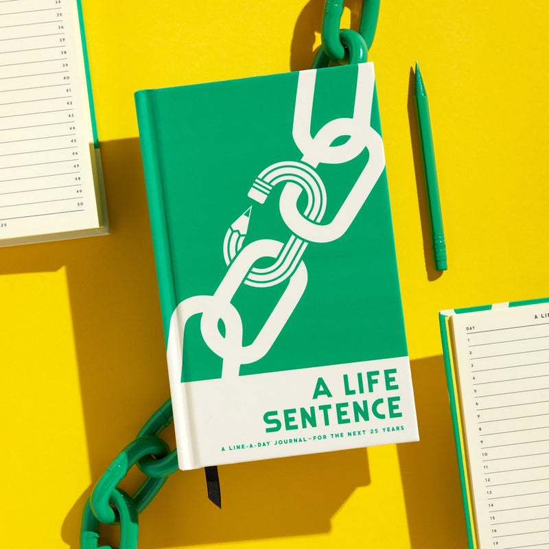 The Brass Monkey A Life Sentence Journal, in green and white with a chain and pencil cover design, is displayed on a yellow background next to a matching pen and two open lined notebooks.
