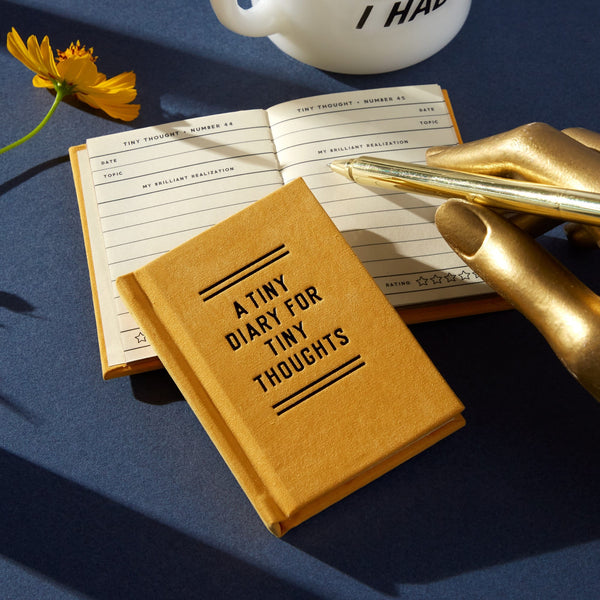The Brass Monkey A Tiny Diary for Tiny Thoughts, a small yellow felt-covered hardbound book, rests on a blue surface next to an open page, with a gold pen holder, pen, yellow flower, and white mug nearby.