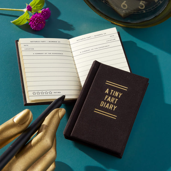 A Tiny Fart Diary by Brass Monkey, a small black mini journal, rests on a teal surface beside an open lined diary. A gold hand sculpture holds a pen, with glimpses of a purple flower and clock in the background.