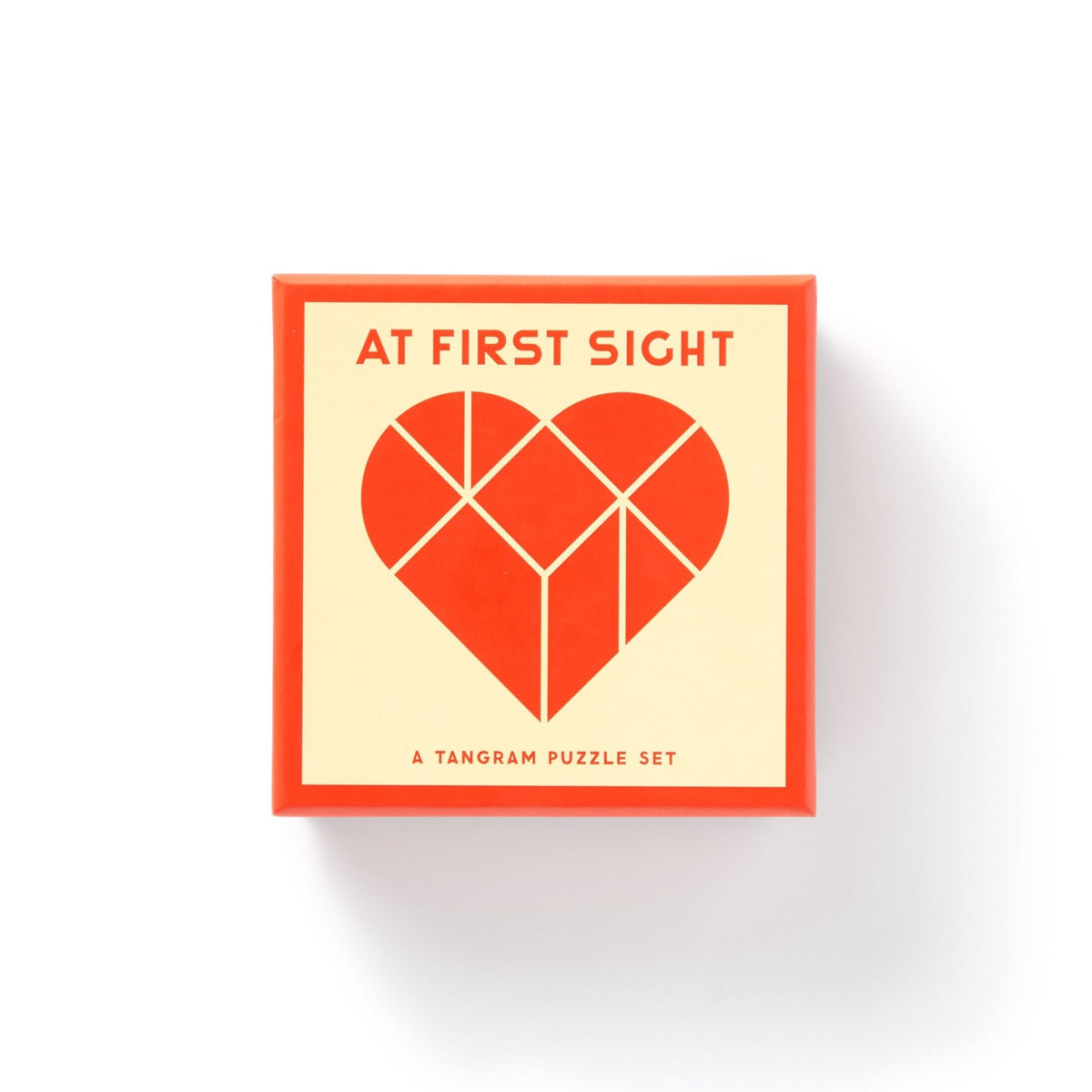 The Brass Monkey At First Sight Tangram Puzzle features a square box with a geometric red heart on the lid, “AT FIRST SIGHT” above it, and “A TANGRAM PUZZLE SET” below. The box has a red border and cream background.