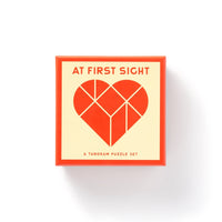 The Brass Monkey At First Sight Tangram Puzzle features a square box with a geometric red heart on the lid, “AT FIRST SIGHT” above it, and “A TANGRAM PUZZLE SET” below. The box has a red border and cream background.