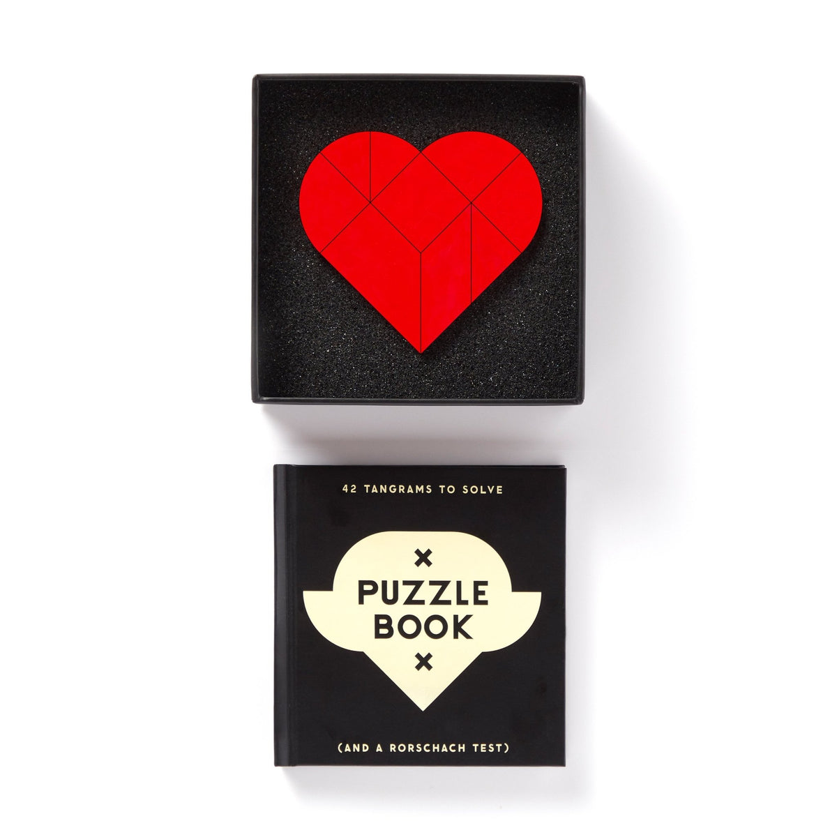 The Brass Monkey At First Sight Tangram Puzzle, featuring a red heart in a black box, sits atop a black puzzle book titled Puzzle Book (and a Rorschach test) for a striking display of unique puzzles against a white background.