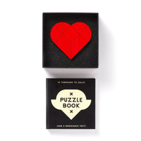 The Brass Monkey At First Sight Tangram Puzzle, featuring a red heart in a black box, sits atop a black puzzle book titled Puzzle Book (and a Rorschach test) for a striking display of unique puzzles against a white background.