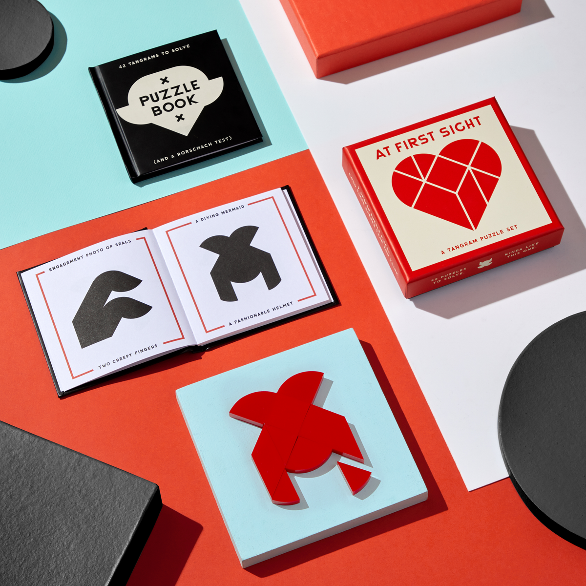 A flat lay of the Brass Monkey At First Sight Tangram Puzzle shows a red heart-shaped puzzle, its matching box, a puzzle book, and open pages with unique challenges, all displayed on vibrant backgrounds.