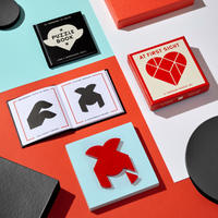 A flat lay of the Brass Monkey At First Sight Tangram Puzzle shows a red heart-shaped puzzle, its matching box, a puzzle book, and open pages with unique challenges, all displayed on vibrant backgrounds.