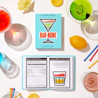 The Brass Monkey Bar None Drink Journal rests on a white surface, open to a Flying Goose recipe page with an illustrated cocktail, surrounded by vibrant drinks, lemons, and garnishes.