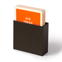 A stack of Because This Dog Is Embarrassing - Pet Shame/Praise Deck cards by Brass Monkey, with an orange ATE TRASH card on top, held upright in a black holder—ideal for capturing your dogs unforgettable milestone moments.