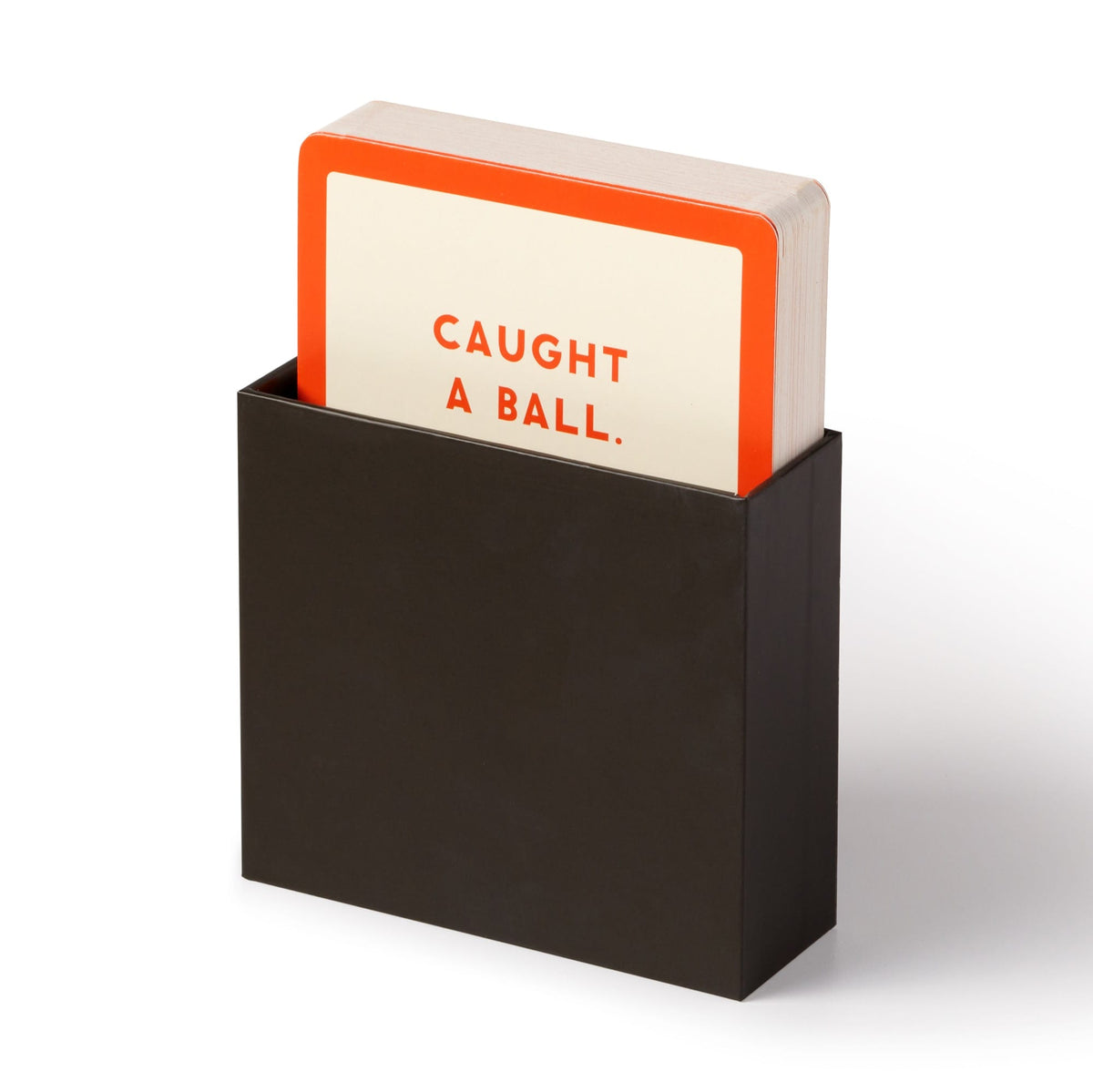 A stack of Because This Dog Is Embarrassing cards by Brass Monkey sits in a black box. The top card, orange-bordered with bold red text on white, reads CAUGHT A BALL.—a fun pick for pet shame/praise dog milestone cards.