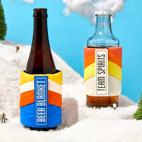 Two bottles in Brass Monkeys Beer Blanket Puffyvest Coozie Set, labeled BEER BLANKET and TEAM SPIRITS, stand on snow with figurines and trees under a blue sky. Made from durable, water-resistant nylon for lasting use.