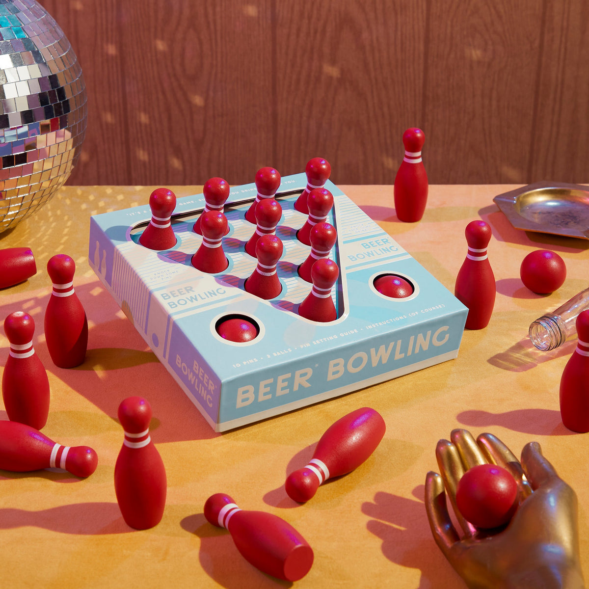 The Brass Monkey Beer Bowling Drinking Game Set features red MDF pins on a yellow surface, a metallic mannequin hand holding a pin, a disco ball, scattered pins, and a bottle for the ultimate fun tabletop bowling experience.