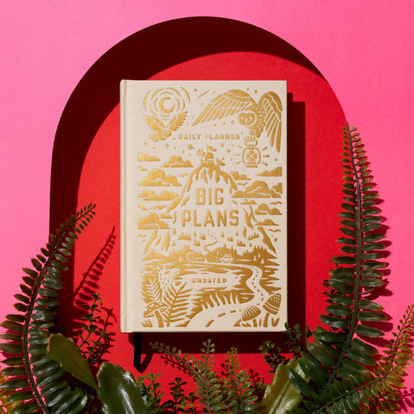The Brass Monkey Big Plans Undated Standard Planner, featuring vintage-inspired beige covers with gold embossed details, is displayed on a red and pink background, accented by green ferns at the bottom.