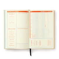 A Big Plans Undated Standard Planner by Brass Monkey opens to a retro two-page January spread: the left page for achievements and notes, the right with a grid calendar, goals, birthdays, and a black ribbon bookmark.