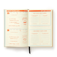 The Big Plans Undated Standard Planner by Brass Monkey features sample daily entries on the left and usage instructions on the right, with orange and blue headings, a black ribbon bookmark, and a vintage-inspired design.