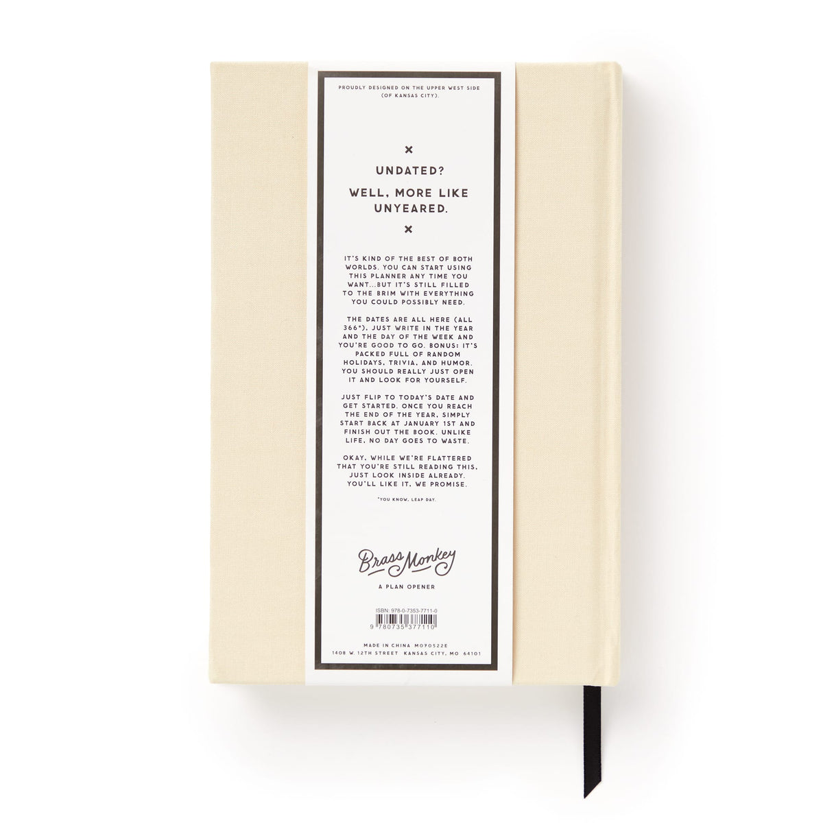 A cream-colored Brass Monkey Big Plans Undated Standard Planner seen from the back, featuring a white label with black text about its flexible design, plus a black ribbon bookmark and vintage-inspired style.