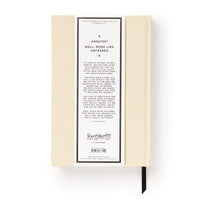 A cream-colored Brass Monkey Big Plans Undated Standard Planner seen from the back, featuring a white label with black text about its flexible design, plus a black ribbon bookmark and vintage-inspired style.