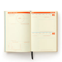 The Big Plans Undated Standard Planner by Brass Monkey features January preview pages with sections for goals, monthly overviews, charts, and planning space for January 1 and 2 in a clean layout with orange highlights and grid lines.