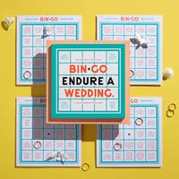 The Brass Monkey Bin-go Endure A Wedding Bingo Book sits atop four bingo cards on a yellow background, surrounded by white flowers, gold rings, and pearl-like beads—perfect for people-watching at weddings.
