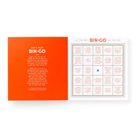 An open Bin-go Get Some Drinks Bingo Book by Brass Monkey shows a bright orange “Let’s Play BIN-GO” page and a bingo card filled with bar and restaurant game phrases—plus an X-marked square for people-watching fun.