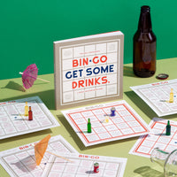 A boxed Brass Monkey Bin-go Get Some Drinks Bingo Book sits on a table with bingo cards, drink umbrellas, game pieces, a bottle cap, and an empty brown glass bottle—perfect for bar or restaurant fun.
