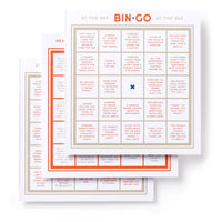 Brass Monkey’s Bin-go Get Some Drinks Bingo Book features three overlapping bingo cards with common bar phrases and scenarios; the center square has a blue X, and “BIN×GO” in red tops the main card—perfect for people-watching.