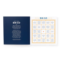 An open Bin-go To A Dumb Meeting Bingo Book by Brass Monkey shows playing instructions on the left page and a bingo card with classic office phrases on the right, with one square marked by an orange X.